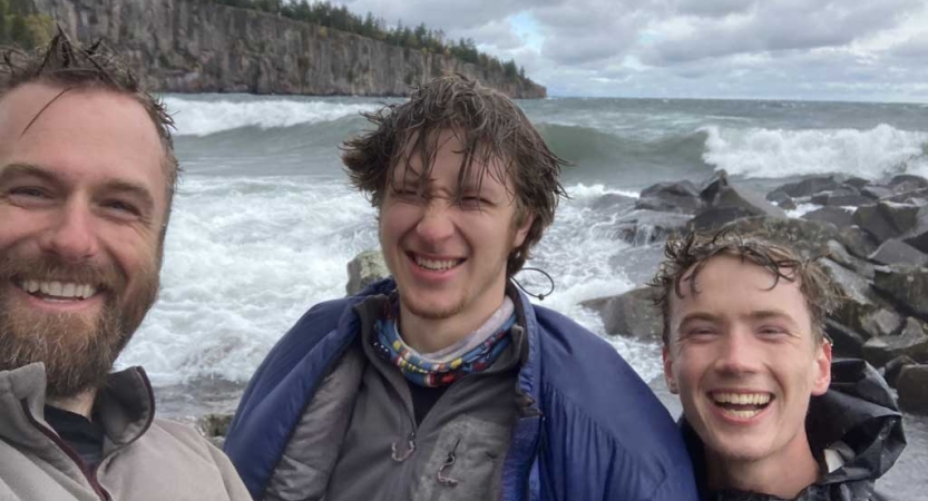 Three people smile standing on the shore of a lake with large waves in the background. 